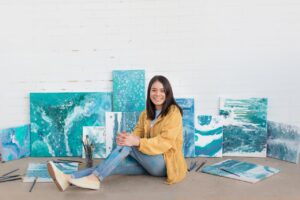 Young female artist sitting with abstract ocean-themed paintings and brushes.